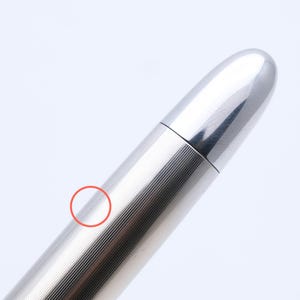 May include: A close-up of a silver pen with a shiny, metallic finish. The pen has a ribbed design on the barrel and a smooth, rounded top. The pen is set against a plain white background.