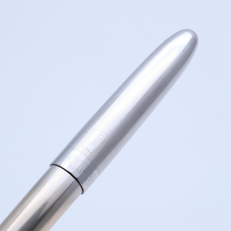 May include: A close-up of a silver pen with a rounded tip. The pen has a textured, ridged design on the barrel and the word "OMAS" is visible. The pen is set against a light blue background.