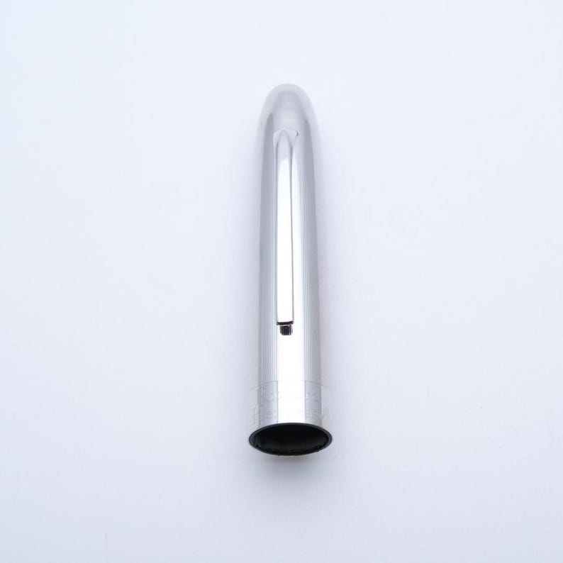 May include: A silver pen cap with a rounded top and a black opening. The pen cap has a clip and vertical lines etched into the metal. The pen cap is on a white background.