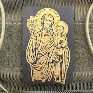 May include: A rectangular patch featuring a gold illustration of Saint Joseph holding the child Jesus. The artwork is set against a dark background, with the figures outlined in gold. The patch is likely designed for attachment to tactical gear.