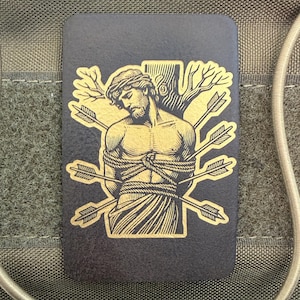 May include: A rectangular, dark gray patch featuring a gold illustration of a man tied to a tree, pierced by arrows. The image is set against a backdrop of olive green fabric and elastic cords.