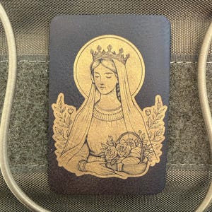 Pode incluir: Remendo retangular com uma ilustração dourada de uma mulher coroada segurando rosas, sobre um fundo escuro. O design inclui um halo e detalhes dourados. Adequado para roupas ou acessórios, provavelmente um remendo decorativo.