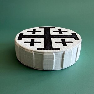 May include: A white, round object with a black Jerusalem cross design on top. The object has a layered appearance, with a series of stacked, thin layers forming the sides. The background is a solid, muted green.