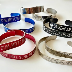 May include: A collection of open cuff bracelets in various colors, including blue, red, black, and silver. Each bracelet features engraved text and a cross symbol. The text includes phrases like "Lo, Here Am I" and "Send Me."