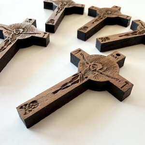 Könnte beinhalten: Mehrere dunkelbraune Holzkreuze mit detaillierten Schnitzereien einer Figur am Kreuz. Die Kreuze tragen die Inschrift "INRI" und ein kleines Loch zum Aufhängen. Sie sind auf einer weißen Oberfläche angeordnet.