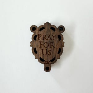 May include: A wooden religious pendant with the words "PRAY FOR US" engraved in the center. The pendant has a decorative, ornate shape with cut-out details and the text "USA" at the bottom. The pendant is a dark brown color.
