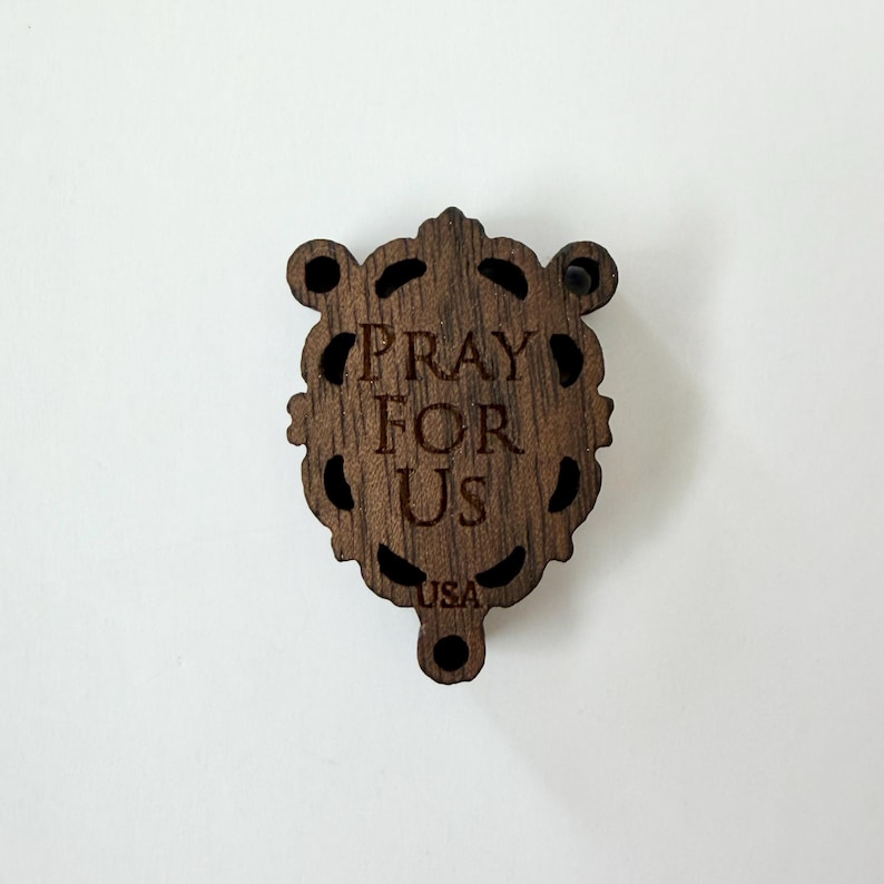 May include: A wooden religious pendant with the words "PRAY FOR US" engraved in the center. The pendant has a decorative, ornate shape with cut-out details and the text "USA" at the bottom. The pendant is a dark brown color.