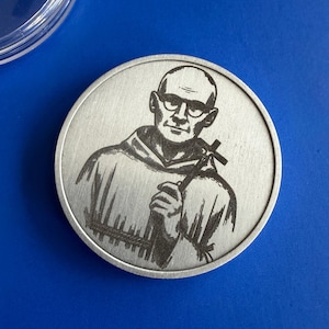 May include: A silver-colored round coin featuring a detailed engraving of a man wearing glasses and a robe, holding a cross. The coin has a brushed metal finish and is set against a blue background.