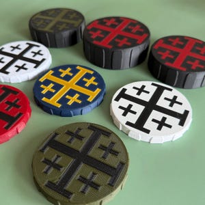 Op de afbeelding: Een verzameling ronde, decoratieve objecten met het ontwerp van het Jeruzalemkruis. De objecten zijn in verschillende kleuren, waaronder rood, zwart, wit, blauw en olijfgroen. Het Jeruzalemkruis is prominent aanwezig op de bovenkant van elk object.