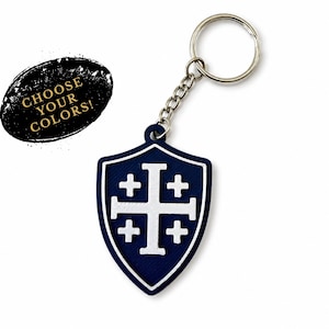 May include: A navy blue shield-shaped keychain featuring a white Jerusalem cross emblem. The keychain includes a silver-toned metal ring and chain. A black oval graphic to the left contains the gold text CHOOSE YOUR COLORS!