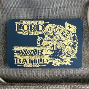 May include: A navy blue rectangular patch with gold text and a detailed illustration of a soldier. The text reads "Blessed be the Lord, My Rock" and "Who trains my hands for war and my fingers for battle. Psalm 144:1."