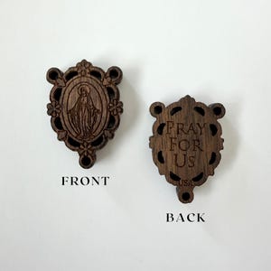May include: Two wooden religious pendants. The front pendant features an engraved image of a religious figure within an oval frame, surrounded by floral accents. The back pendant has the words "PRAY FOR US" and "USA" engraved. Both pendants are dark brown.