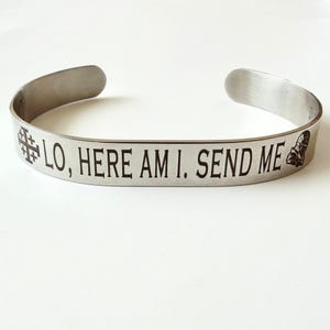 May include: A silver-colored cuff bracelet with the engraved text "LO, HERE AM I. SEND ME." The bracelet features a cross symbol and a small boot design. The open-ended design allows for easy wear.
