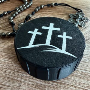 May include: A black, round container with a textured surface, featuring a white graphic of three crosses. A rosary with dark gray beads and a silver crucifix is partially visible in the background. The container is on a wooden surface.