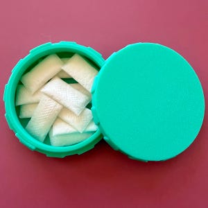 Puede incluir: Un recipiente verde turquesa abierto, que revela varias bolsas rectangulares blancas. El recipiente está sobre una superficie granate. La tapa es redonda y el recipiente tiene un borde texturizado.
