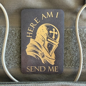 May include: A rectangular, dark gray patch with the words "HERE AM I SEND ME" in gold lettering. The patch features a gold illustration of a knight's helmet with a cross emblem, set against a textured background.