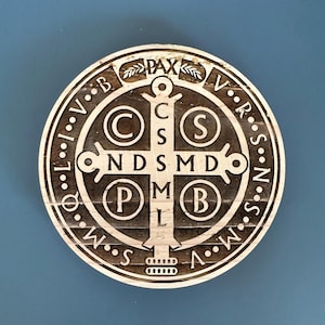 Puede incluir: Medalla redonda de madera de San Benito con un diseño de cruz detallado. El anillo exterior presenta texto y puntos, con la palabra "PAX" en la parte superior. La cruz tiene las letras "C S M L N D S M D B" y otros símbolos. La medalla es de color marrón claro.