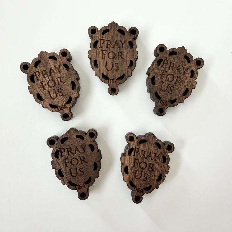May include: Five wooden prayer tokens with a dark brown finish. Each token is shield-shaped with a decorative border and the words "PRAY FOR US" engraved. The word "USA" is also engraved on each token.