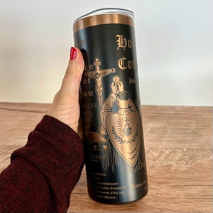May include: A black stainless steel tumbler with a copper-colored top and accents. The tumbler features an engraved religious design with text and imagery of a priest, a crucifix, and other religious symbols.