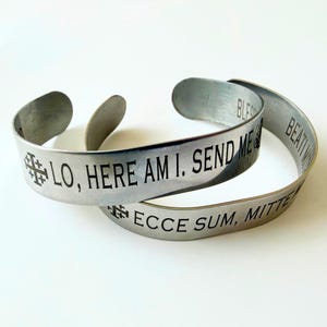 Puede incluir: Dos pulseras de puño de metal plateado con texto grabado y símbolos de cruz. Una pulsera dice "LO, HERE AM I. SEND ME." La otra pulsera dice "ECCE SUM, MITTE ME."