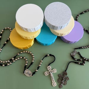 May include: A collection of colorful, round jar lids in white, gray, yellow, blue, and purple. Three rosaries with silver-toned beads and crucifix pendants are also present, set against a green background.