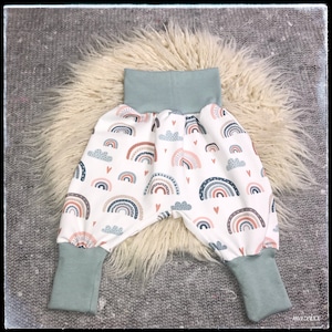 Könnte beinhalten: Babyhose mit einem Muster aus bunten Regenbogen und Wolken auf weißem Grund. Die Hose hat einen hellblauen Bund und Bündchen. Das Design umfasst rosa, blaue und braune Regenbogen. Hergestellt aus weichem Stoff.