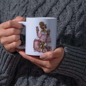 May include: A white ceramic mug featuring a graphic illustration of a leopard character wearing sunglasses and a pink sweatshirt with the word MESSY printed on the front. The character is surrounded by pink shopping bags and stacks of cash.