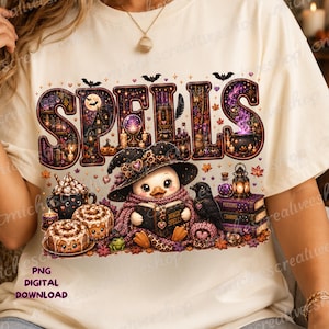 May include: A cream-colored t-shirt featuring the word SPELLS in large, decorative letters filled with magical imagery. Below, a cartoon duck wearing a witch hat and scarf reads a book, surrounded by a crow, cinnamon rolls, and potion bottles.