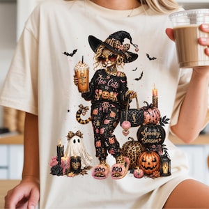 May include: A cream-colored t-shirt featuring a graphic of a leopard-print witch wearing sunglasses and a hat, holding a coffee cup. The text reads Too Cute to SPOOK and SPOOKY SEASON. The design includes pumpkins, a ghost, and candles.
