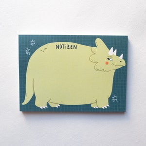 Könnte beinhalten: Notizblock mit einem Cartoon-Triceratops in Hellgrün mit weißen Hörnern und einem lächelnden Gesichtsausdruck. Der Notizblock hat einen teal-farbenen Gitterhintergrund mit dem Wort "NOTIZEN" in Schwarz. Weiße Sterne sind gezeichnet.