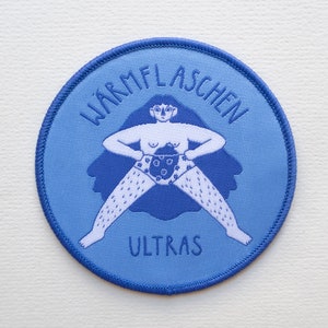 Könnte beinhalten: Blauer gestickter Aufnäher mit einer Cartoon-Illustration einer Person, die eine Wärmflasche hält. Der Text "Wärmflaschen Ultras" ist um die Person herum gestickt.