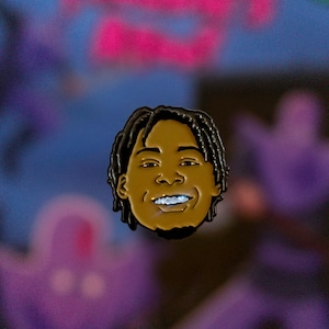May include: A pin featuring a smiling face with dark hair and a brown complexion. The pin has a gold outline and is set against a blurred background with pink and purple hues. The pin is a portrait of a person.