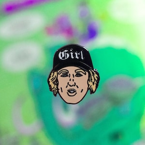 Könnte beinhalten: Eine schwarz-weiße Emaille-Anstecknadel mit einer Cartoon-Illustration einer Person, die eine schwarze Baseballkappe mit dem Wort "Girl" in Weiß auf der Vorderseite der Kappe trägt.
