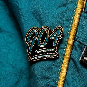 May include: A gold-edged pin with the number "904" in black and teal lettering. The pin is set against a teal fabric background with a gold zipper and a black and white tag.