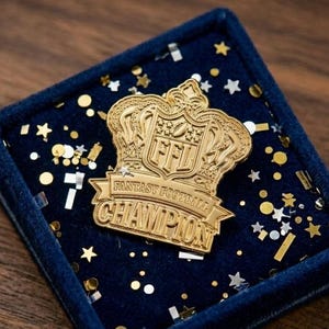 May include: A gold-colored Fantasy Football Champion pin, shaped like a crown, rests on a navy blue velvet surface. The pin features the letters "FFL" and the words "Fantasy Football Champion". The background is decorated with gold and silver confetti.