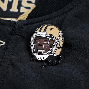 May include: A gold and black enamel pin featuring a football player's helmet with a smiling face. The pin is set against a black jacket with gold accents and the word "SAINTS" visible.