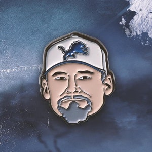May include: A blue and white enamel pin of a man wearing a baseball cap with a lion logo. The man has a beard and mustache.
