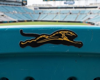 Può includere: Primo piano di un sedile dello stadio turchese con un logo di giaguaro nero e oro. Il logo è un'immagine stilizzata di un giaguaro in movimento. Lo sfondo mostra uno stadio con sedili turchesi e un grande tabellone segnapunti.