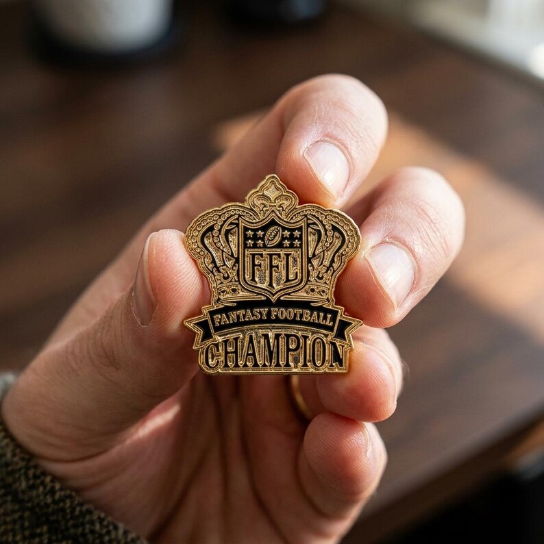 Op de afbeelding: Een goudkleurige Fantasy Football Champion-speld met een kroonontwerp. De speld heeft de letters "FFL" in een schild en de woorden "Fantasy Football Champion" op een banner.