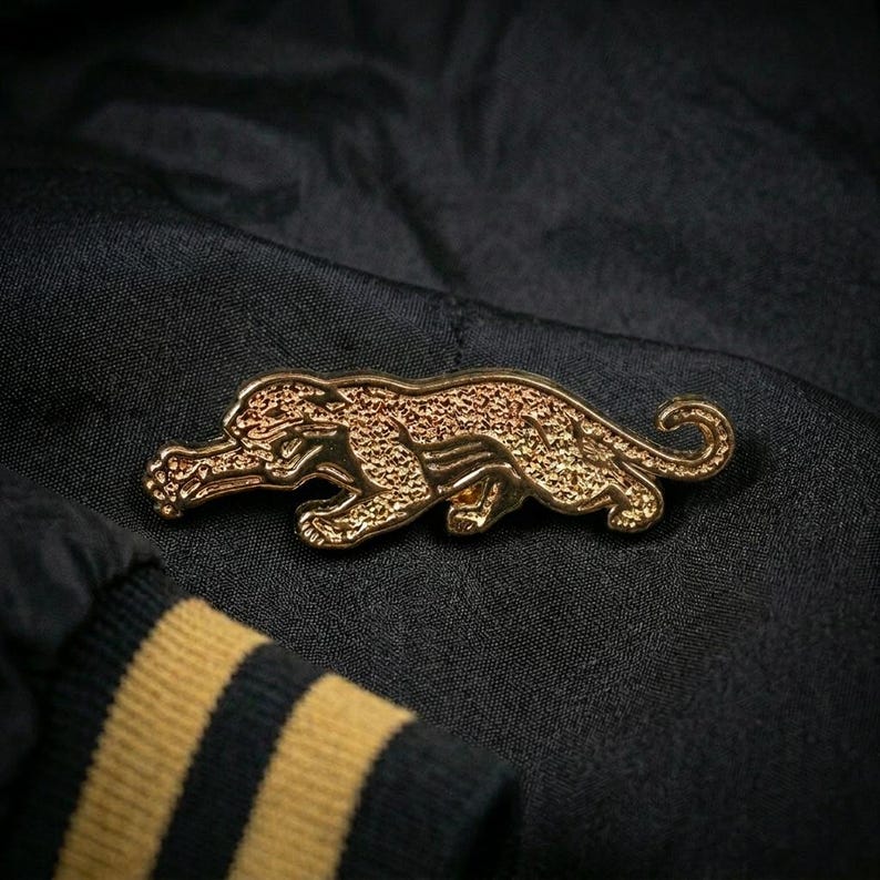 May include: A gold-colored panther pin with a textured surface. The pin is in a crouching position, with its tail curled. The pin is set against a black fabric background, with a sleeve featuring yellow stripes.