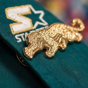 May include: A gold-colored pin in the shape of a jaguar, attached to a dark green fabric. The pin is highly detailed, with textured surfaces. The fabric has a white and gold embroidered logo.