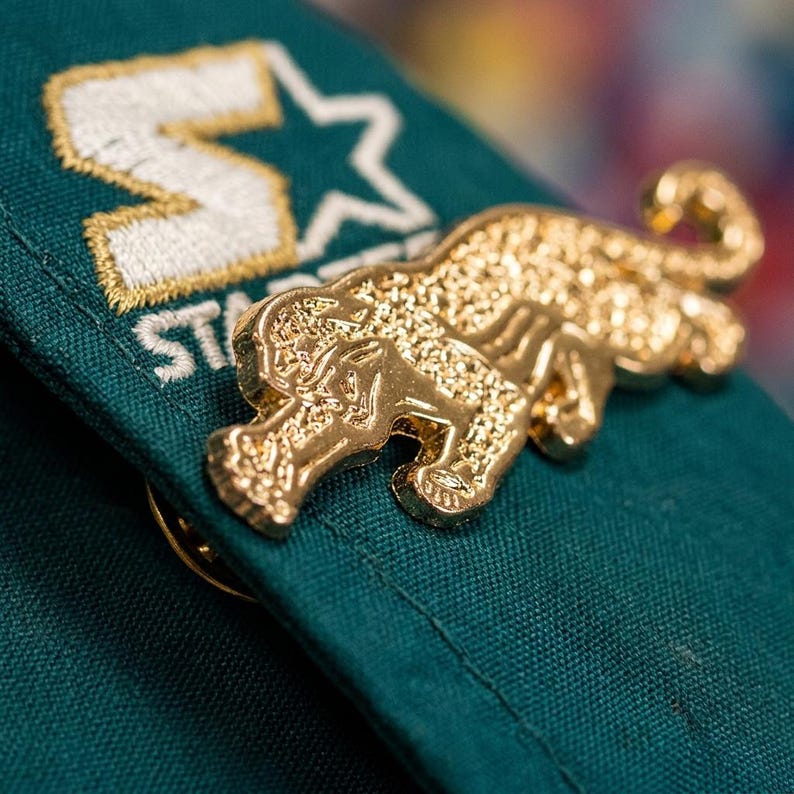 May include: A gold-colored pin in the shape of a jaguar, attached to a dark green fabric. The pin is highly detailed, with textured surfaces. The fabric has a white and gold embroidered logo.