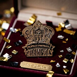 Op de afbeelding: Een goudkleurige Fantasy Football Champion-speld met een kroonontwerp, geplaatst in een bordeauxrode fluwelen doos. De speld toont het NFL-logo en de woorden "Fantasy Football Champion". Een klein plaatje leest "1st Place".
