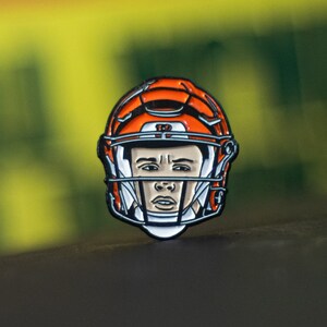 May include: Orange and black football player pin with a white face and a black helmet with a white number 17 on it.