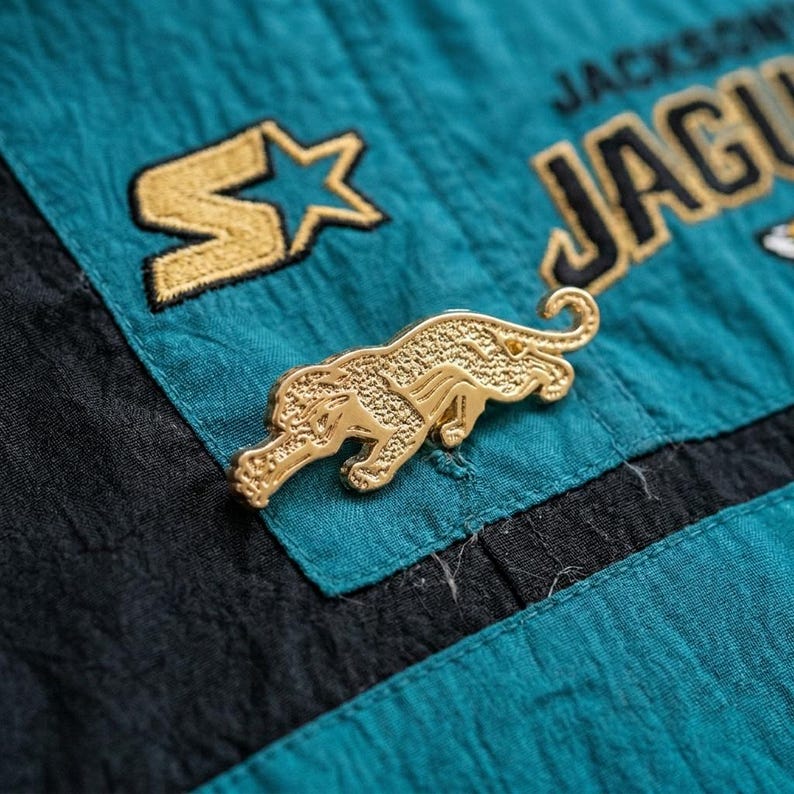May include: A gold-colored pin in the shape of a jaguar, set against a teal and black fabric background. The fabric has gold embroidered lettering that reads "JACKSONVILLE JAGUARS" and a gold star logo. The pin is detailed with the jaguar's features.