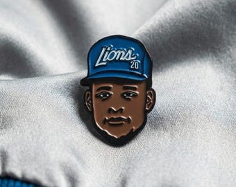 Può includere: Una spilla con un'illustrazione stilizzata di una persona che indossa un berretto da baseball blu con la scritta "Lions 20". La spilla ha un bordo nero e un viso marrone.