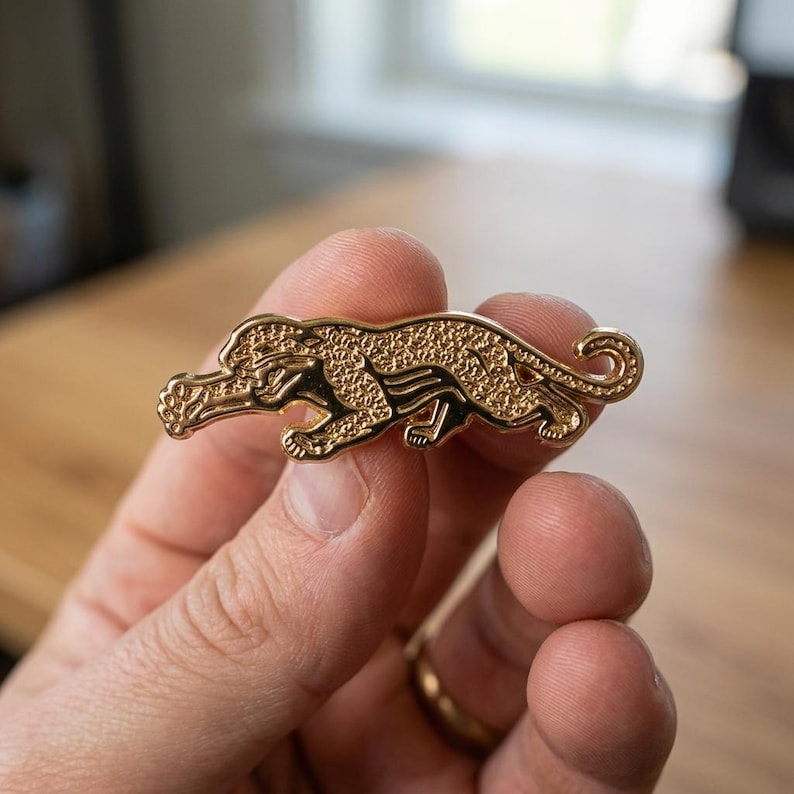 May include: A gold-colored pin in the shape of a jaguar. The pin has intricate detailing, with a textured surface and black accents. The jaguar is depicted in a crouching position, with its tail curled. The pin is held in a person's hand.