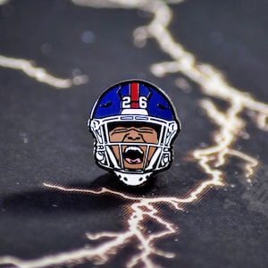 May include: A blue and white football helmet with a player's face and the number "26" on the front. The helmet is a pin.