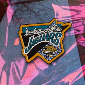 Peut inclure: Une épingle de logo de l'équipe des Jaguars de Jacksonville. L'épingle présente le logo de l'équipe en bleu sarcelle, noir, or et blanc. Le logo comprend le nom de l'équipe et une tête de jaguar.
