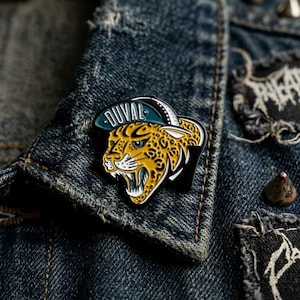May include: A yellow and black jaguar enamel pin with the word "DUVAL" on a teal hat. The pin is set against a blue denim jacket.